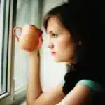 Femme fatiguée à la fenêtre avec une tasse, le matin : la fatigue persistante ne vient pas toujours d un manque de vitamines