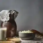 Photographie d’illustration : verre de lait, fromage frais dans un bol, pichet en terre et œuf sur une table en bois