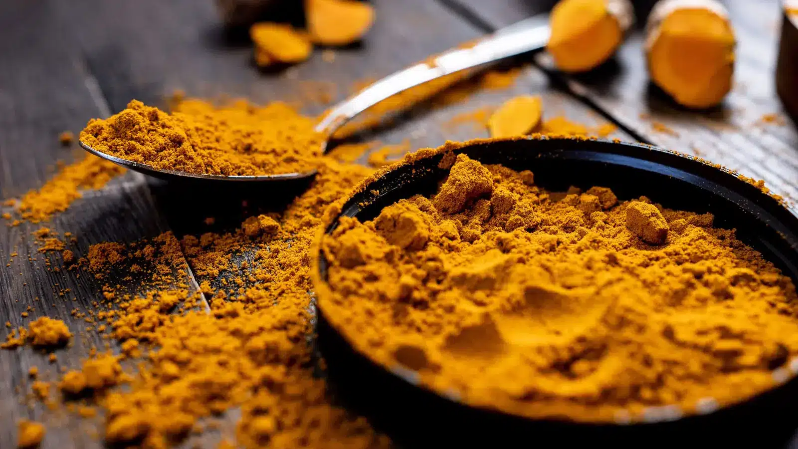 Photographie d’illustration : poudre de curcuma dans une assiette noire et une cuillère, sur une table en bois