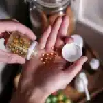 يد رجل تحمل كبسولات مكمّل غذائي فوق طاولة خشبية، ضوء طبيعي