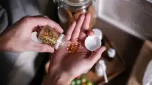 يد رجل تحمل كبسولات مكمّل غذائي فوق طاولة خشبية، ضوء طبيعي